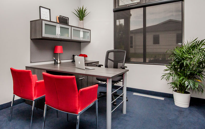 Innspace executive office. L-shaped desk with black chair and 2 modular red arm chairs on opposite side. Red lamp, thumb tac area and overhead cubbies on wall side of desk. Plant, blue carpeting and large window with black sunshade.