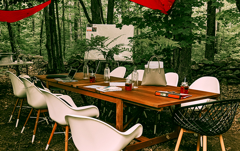 Boardroom table with white chairs in an outdoor forrest setting. Glasses of iced tea on the table, a purse, books and an open lap top.