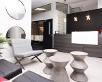 Innspace entry and waiting area. Black and Gray modern seating, decorative pillows, plant, round large mirrors on wall, 3 small decorartive coffee tables, large reception desk with pendant lighting, red roses in vase, innspace logo in black on black wall.
