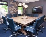 The Quarry Boardroom. Large and square wood conference table, 10 blue modern swivel chairs around it, 3 globe style pendant lights hanging from ceiling, lazy susan and water glasses, wall-mounted smart TV, brick design accent wall, plant and 2 large windows.