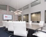 The Ridge Room Boardroom. Long room with vaulted ceiling, boardroom table and 10 white modern swivel chairs. 2 large rectangle shaped pendant lights hanging from ceiling, wall-mounted smart TV displaying innspace logo, square and rectangle line design done in red on TV wall.
